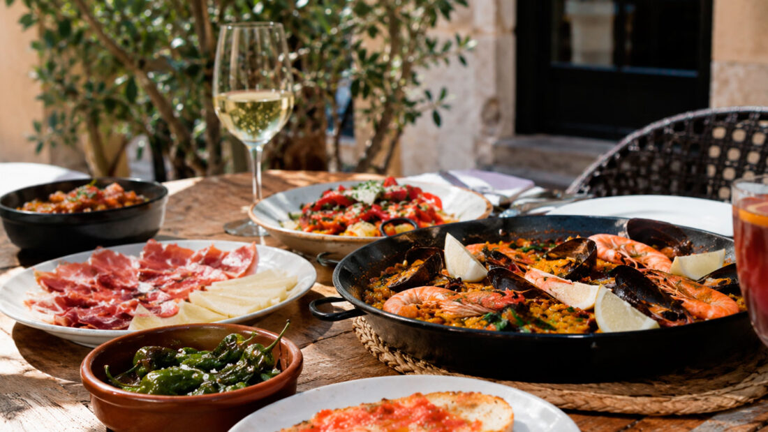 Traditional Catalan and Spanish dishes on a restaurant terrace in Barcelona, including seafood paella, pa amb tomàquet, padrón peppers and cured ham.
