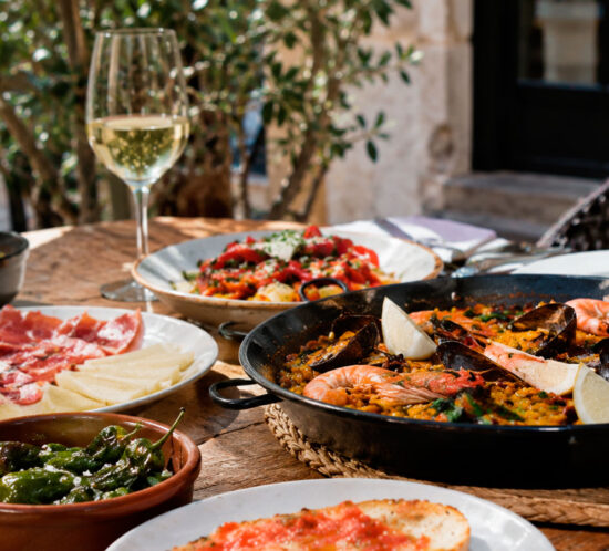 Traditional Catalan and Spanish dishes on a restaurant terrace in Barcelona, including seafood paella, pa amb tomàquet, padrón peppers and cured ham.